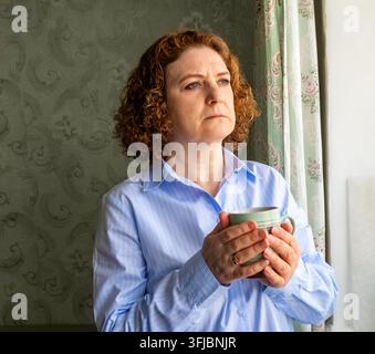Reife Frau hält eine Tasse und schaut nachdenklich aus dem Fenster in einem häuslichen Porträt Stockfoto