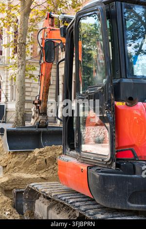 Ein kleiner orangefarbener Raupenbagger, der bei Grabungsarbeiten während der Straßenreparatur und der Versorgungseinrichtung auf einer Stadtstraße arbeitet. Stockfoto