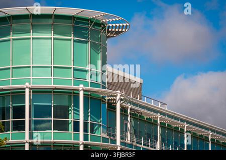 Modernes Glas-Bürogebäude mit einer gebogenen, grün getönten Fassade und Stahlsäulen, das Himmel und Wolken reflektiert. Corporate Business Architektur vermittelt p Stockfoto