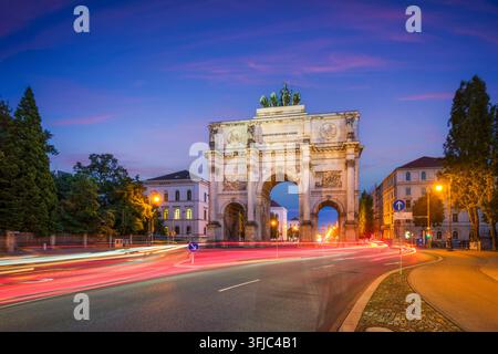 Dramatische Abendansicht des Siegestors in München, Bayern, Deutschland, mit lebendigen roten und weißen Lichtspuren von Autos auf der Straße. Stockfoto
