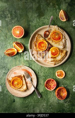 Hausgemachte Crêpes-Pfannkuchen serviert auf Keramiktellern mit blutigen Orangen und Rosmarin-Sirup mit geschnittenen sizilianischen roten Orangen über grünem Strukturhintergrund. Oberansicht, Platz Stockfoto
