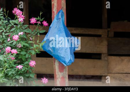 Ein blauer Plastikbeutel, der an einem Holzpfosten neben einem rosa Blumenstrauch in einer rustikalen Veranda-Einstellung mit ruhiger ländlicher Atmosphäre und verwitterter Struktur hängt Stockfoto