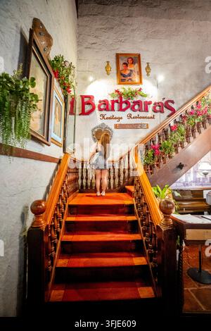 Eine weibliche Reisende steht auf dem rot ausgelegten Treppenabsatz eines historischen Kolonialrestaurants in Intramuros und fängt eine klassische kulturelle Reise ein Stockfoto