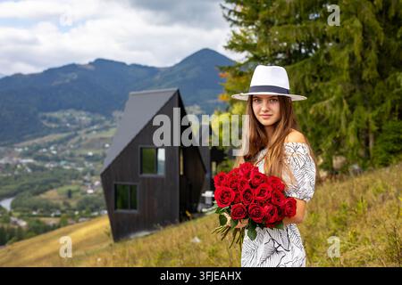 Junge Frau hält rote Rosen in einer Berglandschaft. Junge Frau mit weißem Hut und gemustertem Kleid hält einen Strauß roter Rosen im Freien. Stockfoto
