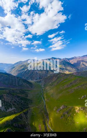 Eine Luftperspektive zeigt ein weites Bergtal in Kirgisistan. Ein gewundener Fluss fließt durch üppiges grünes Gras und felsiges Gelände, umgeben von steilen Stockfoto