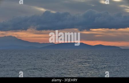 Dramatischer Sonnenuntergang über den Bergen von Evia, gesehen von Artemida, Ost-Attika, Griechenland, mit dunklen Wolken und einem warmen orangefarbenen Horizont. Stockfoto