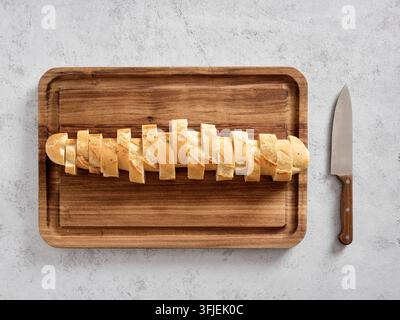 Frisch gebackenes Baguettebrot in kleinere Stücke geschnitten, das auf einem rustikalen Holzbrett mit einem Messer daneben liegt Stockfoto