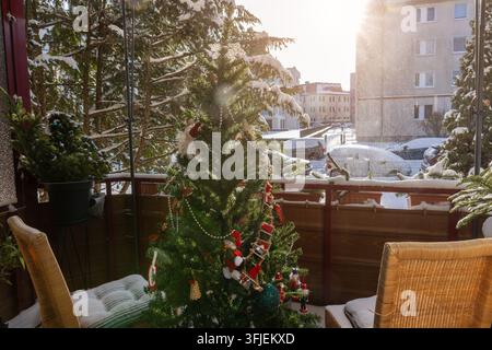Schneebedeckter Weihnachtsmorgen mit Blick auf einen gemütlichen Balkon, geschmückter Tanne, Weidenstühlen und warmem Sonnenlicht. Frischer Schnee bedeckt das Geländer, die Dächer und Stockfoto