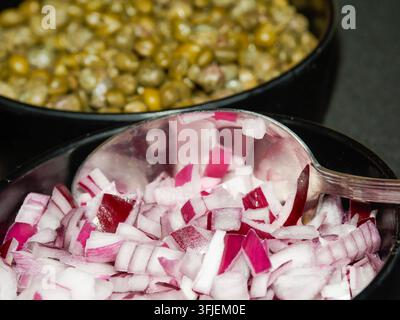 Nahaufnahme von gehackten roten Zwiebeln und grünen Erbsen in schwarzen Schüsseln mit einem Metalllöffel. Stockfoto