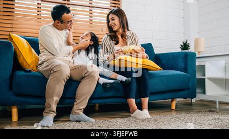 In ihrem gemütlichen, gerillten Haus genießt eine fröhliche Familie Fernsehen mit Popcorn auf dem Sofa. Vater, Mutter, Tochter und Geschwister teilen Gelächter, Lächeln und Momente der Zusammengehörigkeit Stockfoto