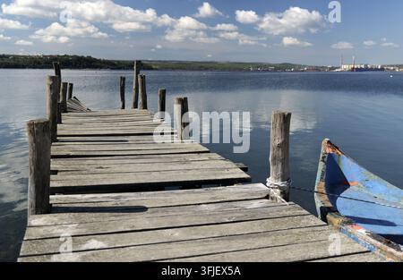 Verwitterter Holzsteg, der sich über ruhiges blaues Wasser erstreckt, mit einem kleinen blauen Fischerboot, das an einem Ankerpfahl festgemacht ist, und zeigt eine friedliche Wassermenge unter einem bri Stockfoto
