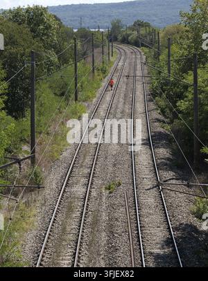 Person in einem orangefarbenen Hemd geht entlang gewundener Bahngleise durch üppige grüne Landschaft, Stromleitungen über Kopf, Hügel und ein See am fernen Horizont Stockfoto