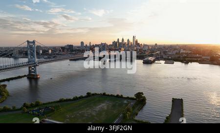 Luftaufnahme rückwärts der Skyline von Philadelphia, Fluss und berühmter Brücke bei Sonnenuntergang. Blick von der Uferlinie der Stadt Camden. Weitwinkelaufnahme steigend. Marinasteg und s Stockfoto