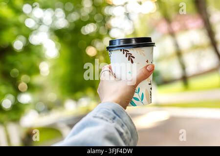 Nahaufnahme einer Frauenhand, die draußen eine Papierkaffeetasse mit schwarzem Deckel hält. Heißes Getränk zum Mitnehmen in einem wunderschönen Sommerpark mit grünen Bäumen und s Stockfoto