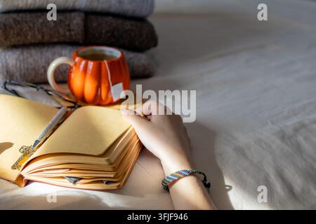 Hand ruht auf offener Tagebuchseite mit Kürbistasse und Pullovern im Hintergrund. Konzept einer ruhigen Tagebuchpause während eines gemütlichen Herbstmorgens Stockfoto