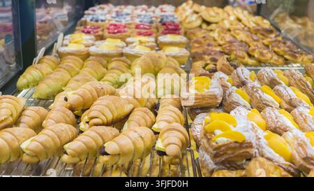 Verschiedene frisch gebackene Croissants, mit Sahne gefüllte Gebäckstücke und mit Obst besetzte dänische Gebäckstücke, die in einer Bäckerei ausgestellt sind und eine Vielzahl von goldbraunen Sorten zeigen Stockfoto