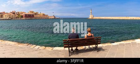 Panoramic view of the Chania waterfront and lighthouse across the sea with people sitting on a bench, Crete, Greece. Stockfoto