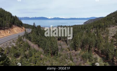 Luftbild des Lake Tahoe in Nevada mit tiefblauem See, schneebedeckten Sierra-Nevada-Bergen, sich windender zweispuriger Autobahn durch dichten Kiefernwald und felsigem Ufer Stockfoto