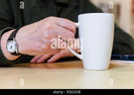 Nahaufnahme der Hand einer reifen Frau, die den Griff einer schlichten weißen Keramiktasse auf einer hellen Holzoberfläche greift, mit klassischer analoger Armbanduhr mit br Stockfoto