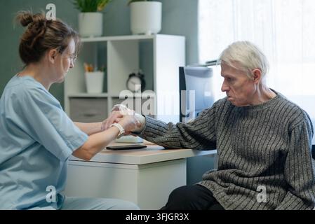 Krankenschwester tröstet kranken älteren Mann mit Handstütze in der Klinik. Gesundheitsfachkraft bietet einfühlsame Betreuung und emotionale Verbindung zu einem Seniorenpatienten mit verletztem Arm und Gipsverband im Innenbereich Stockfoto