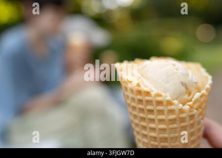 Eine verlockende knusprige Waffeltasche hält eine erfrischende Kugel Vanilleeis vor einem sonnigen Hintergrund im Freien. Dieser appetitliche Sommergenuss sieht bereit zum Essen aus in einer verschwommenen Parkumgebung. Stockfoto