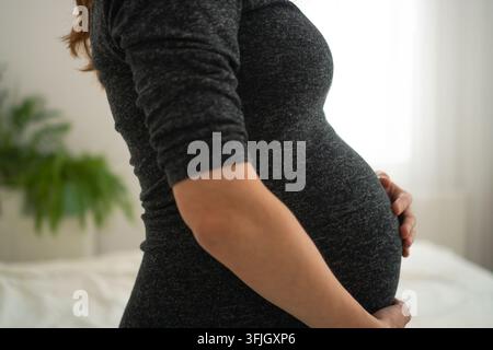 Nahaufnahme des Profils einer schwangeren Frau, die ihren Babybauch sanft in einem ruhigen Schlafzimmer hält. Erwartende Mutter wiegt den Bauch in zärtlicher Seitenansicht. zukünftige Mutter berührt den Bauch mit intimer Fürsorge neben einem weißen Bett Stockfoto