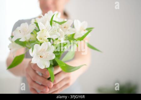 Eine Frau hält mit ausgestreckten Händen einen weißen Blumenstrauß in Richtung Kamera in einer Nahaufnahme. Die Hände, die Blüten anbieten, präsentieren einen zarten, ruhigen Moment des sanften Schenkens in einem hellen Studio. Stockfoto