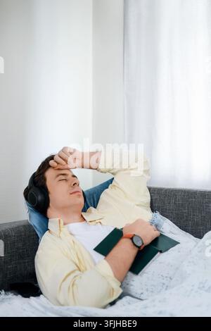 Junger Mann entspannt sich auf dem Sofa mit geschlossenen Augen, hört ein Hörbuch oder Musik in drahtlosen Kopfhörern, ruht sich aus und hält ein Buch zur Entspannung, mental er Stockfoto