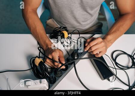 Hochwinkelaufnahme einer Person, die unordentliche Kabel und Adapter an einem Arbeitsplatz verwaltet. Konzept von Technologieeinrichtung, Kabelmanagement, Stromverbrauch Stockfoto