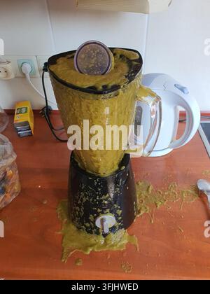 Ein Mixer liegt auf der Seite auf einer hölzernen Küchentheke, bedeckt von und umgeben von verschütteter dicker gelblich-grüner Mixgetränkemischung, die hat Stockfoto