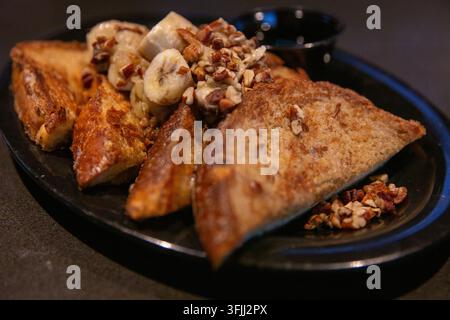 Eine Nahaufnahme, horizontal aufgenommen, zeigt eine schräge Ansicht von dicken goldbraunen French-Toast-Dreiecken, die mit geschnittenen Bananen und zerkleinerten Pekannüssen belegt sind Stockfoto