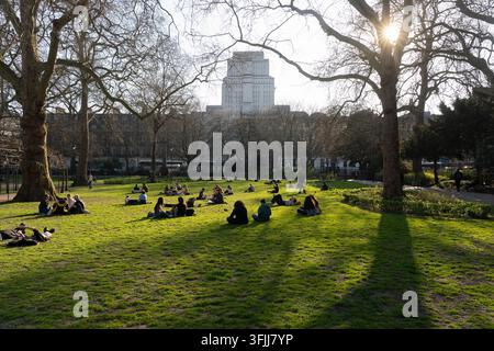 Menschen entspannen sich und knüpfen Kontakte auf dem sonnigen Rasen des Russell Square in London, während das ikonische Senate House im Hintergrund aufragt. Stockfoto