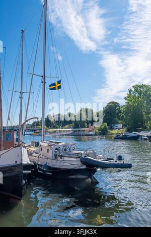 klassisches Segelboot mit einer schwedischen Flagge, die am Takelwerk weht, vertäut neben anderen Schiffen in einem Stockholmer Hafen unter einem strahlend blauen Himmel mit flauschigen Wolken Stockfoto