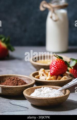 Zutaten für das Kochen von Schokoladen-Mug-Kuchen. Mehl, Kakaopulver, Zucker, Karamell in Holzschüsseln, Milch, Erdbeeren und Minze serviert mit Löffeln und Tasse über grauem Küchentisch. Nahaufnahme Stockfoto