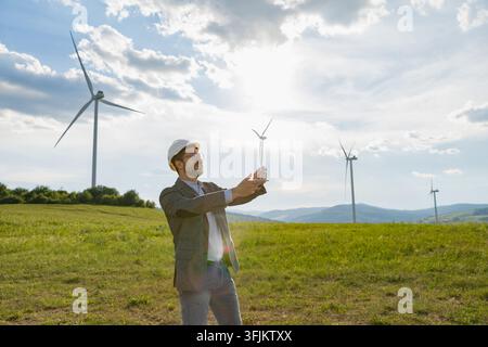 Ingenieur hält ein Windturbinenmodell mit Windmühlen im Hintergrund an einem sonnigen Tag Stockfoto