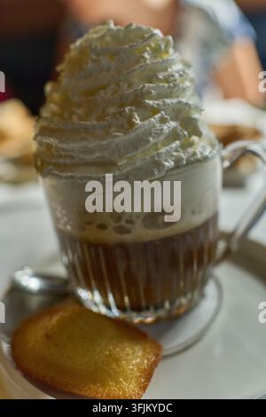 Eiskaffee mit einer großzügigen Sahnehaube, serviert neben einem klassischen Madeleine-Keks auf einem weißen Teller Stockfoto
