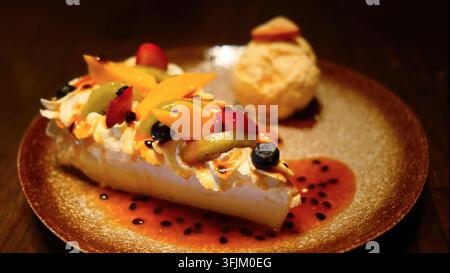 Exquisite Pavlova-Dessertscheibe, belegt mit frischem Mango, Erdbeere, Kiwi und Blaubeeren. Serviert mit einer Kugel Vanilleeis und beträufelt mit Stockfoto