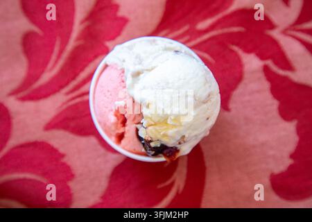 Ansicht von oben: zwei Kugeln rosa und weiße Eiscreme in einer kleinen Schüssel auf lebhaft rotem Blumenstoff mit weicher Unschärfe, cremiger Textur und verspielter Sommerlichkeit Stockfoto