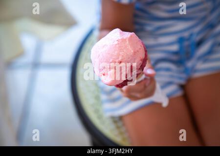 Kind hält eine rosa Eiswaffel nah an die Kamera mit weichem Sommer-Hintergrund und hellen, verspielten Farben, zeigt die cremige Desserttextur und Stockfoto