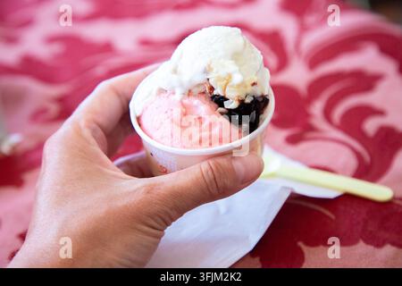 Hand, die eine kleine Tasse mit bunten Eisbechern mit cremiger Topping, dunkler Sauce und rosa Dessert hält, serviert über rotem gemustertem Tuch mit Serviette und Stockfoto