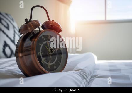 Nahaufnahme einer antiken Weckuhr, die auf dem weißen Bett im Schlafzimmer steht, während das weiche Morgenlicht durch das Seitenfenster scheint. Stockfoto