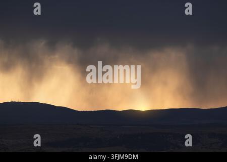 Dunkle Wolken schweben über den Bergen, während ein warmes Sonnenuntergangslicht durchbricht. Ein dramatischer Kontrast zeichnet den Abend aus. Stockfoto