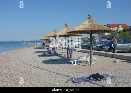 Pirovac, Kroatien, 16. August 2026 Entspannte Strandszene mit Strohsonnenschirmen, kiesiger Küstenlinie, Schwimmern im klaren blauen Wasser, geparkten Autos in der Nähe und Bri Stockfoto