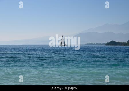 Segelboot segelt über einen blauen Ozean mit Bergschatten im Hintergrund und schafft eine friedliche Küstenszene für Reise- und Lifestyle-Inhalte Stockfoto