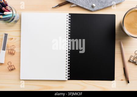 Oberansicht eines offenen Spiralnotizbuch-Mockups mit schwarzen und weißen Seiten auf hellem Holzschreibtisch mit Kaffee und Schreibwaren mit Kopierraum Stockfoto