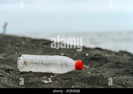 Verlassene Plastikflasche auf einem dunklen Sandstrand. Benutzte Wasserflasche aus Plastik mit rotem Verschluss, halb vergraben im dunklen Vulkansand eines Strandes, mit verschwommenem Hintergrund aus Ozean und entfernten Strandbesuchern. Stockfoto