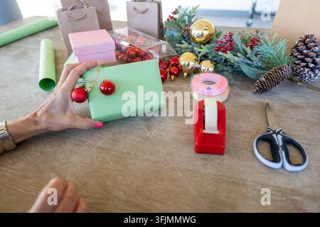 Die Hand einer Person dekoriert eine hellgrüne Geschenkschachtel mit kleinen roten Verzierungen auf einem Holztisch Stockfoto