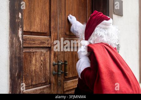Der Weihnachtsmann von hinten gesehen, der an eine große Holztür klopft, während er einen roten Sack voller Weihnachtsgeschenke während der Feiertagsauslieferungen trägt. Stockfoto