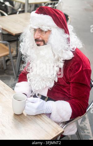 Nahaufnahme-Porträt eines Mannes, der als Weihnachtsmann verkleidet ist und während der Weihnachtszeit mit einer Tasse Kaffee in einem Straßencafé sitzt Stockfoto