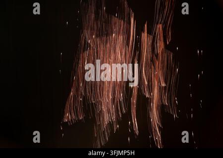 Langzeitbelichtung fängt goldene Funken ein, die in streifigen Bahnen gegen einen schwarzen Hintergrund fallen und ein abstraktes feuriges Lichtmuster erzeugen. Stockfoto